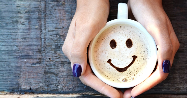 Female holding a cup of coffee with smiley face drawn in foam which may lower cancer risk.