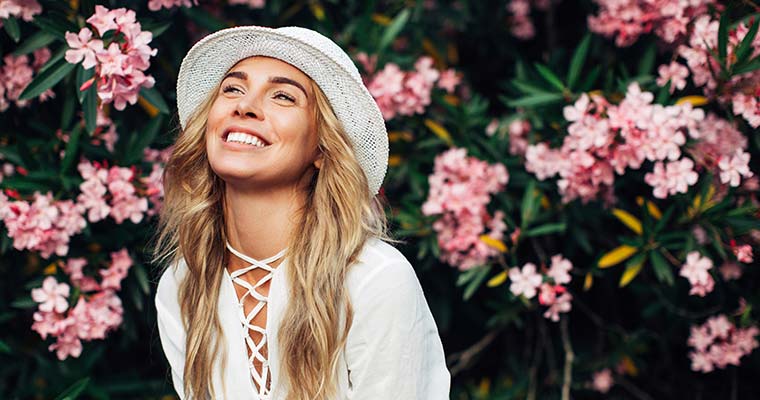 Young blond girl smiling wearing white hat and shirt against flower background, showing off beautiful professional teeth whitening results